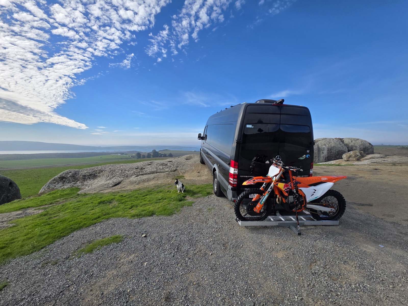 Sprinter van with a dirt bike loaded on the back, parked overlooking the coast — the van this whole project was built for